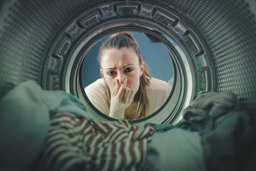 A woman holding her nose while looking at clothes in a washing machine.