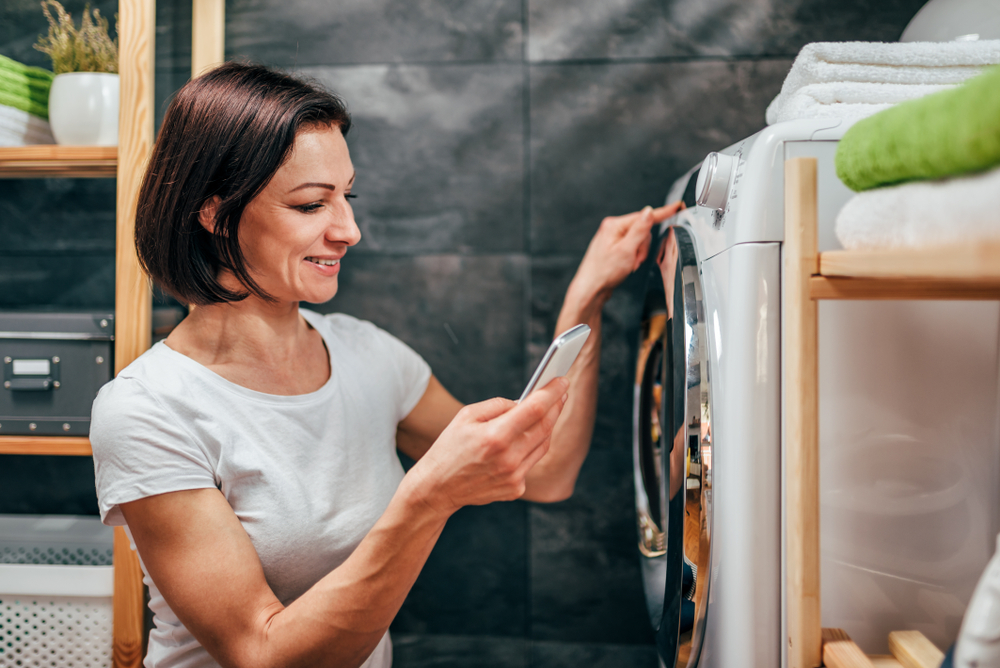 Woman wearing white shirt using smart phone to control washing machine at laundry room